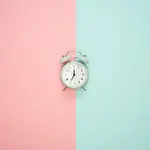 Waiting after your egg retrieval for fertilization & embryo development results can be stressful. Image of clock lying on a surface which is half baby blue and half baby pink.